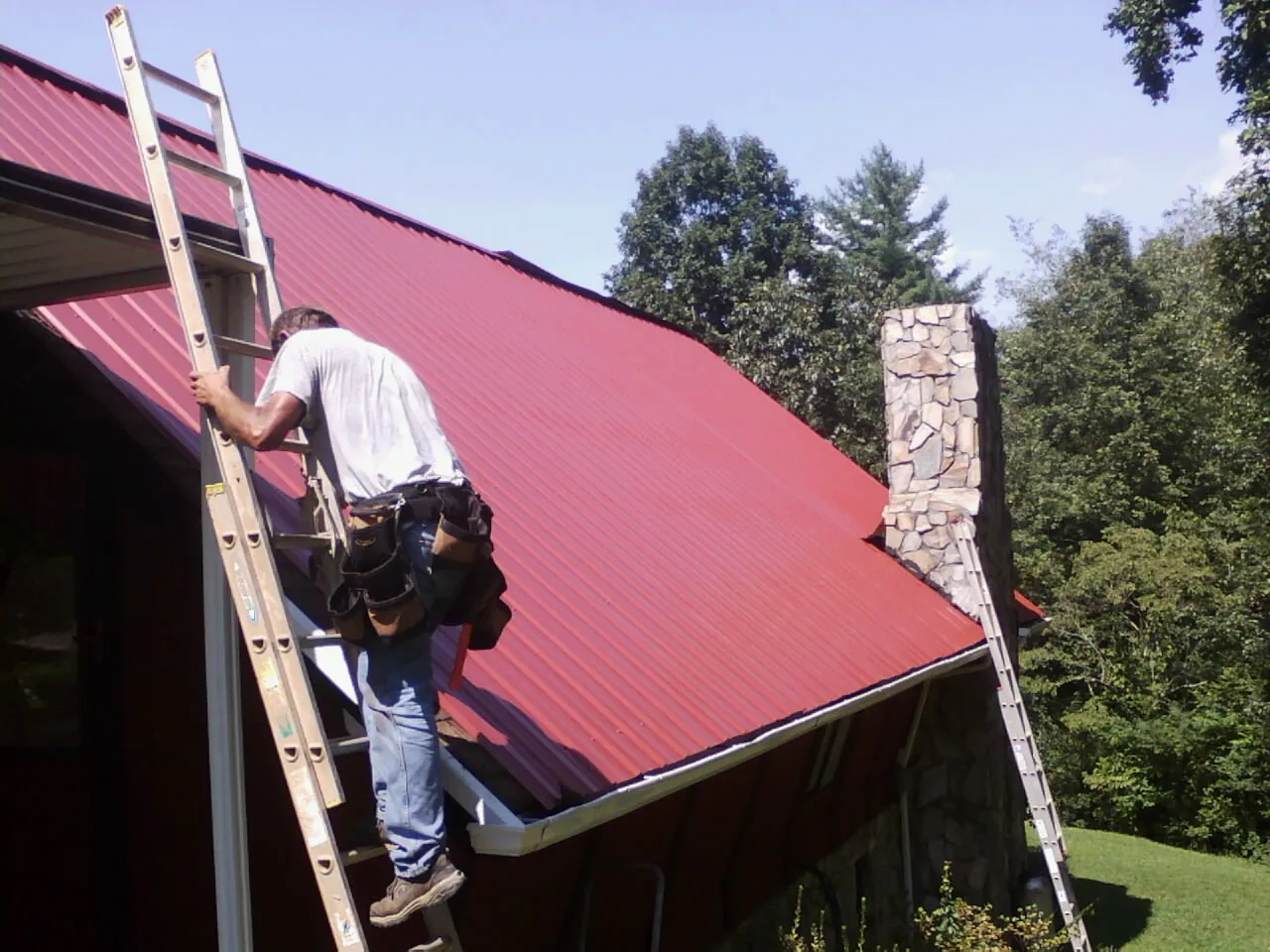 Roofer on a ladder beside a red metal roof and stone chimney — Soffit & Fascia Repair by Roofing Brothers in Belpre